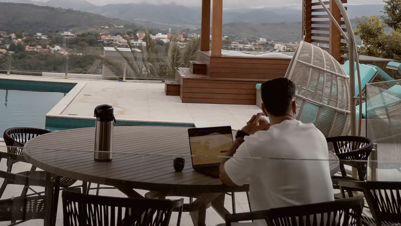Mesa de trabalho ao ar livre pela manhã: notebook aberto, xícara de café e garrafa térmica, com a paisagem ainda em luz baixa